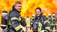 Ein Feuerwehrmann und eine Feuerwehrfrau stehen im Vordergund, dahinter weitere Feuerwehrleute vor hohen Flammen