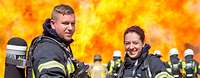 Feuerwehrfrau und Feuerwehrmann Ein Feuerwehrmann und eine Feuerwehrfrau stehen im Vordergund, dahinter weitere Feuerwehrleute vor hohen Flammen
