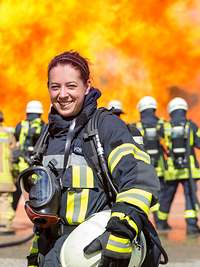 Feuerwehrfrau und Feuerwehrmann Ein Feuerwehrmann und eine Feuerwehrfrau stehen im Vordergund, dahinter weitere Feuerwehrleute vor hohen Flammen