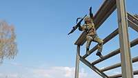 Ein Soldat in Uniform spring mit seinem Gewehr von der Plattform des Sprunggerüstes auf der Hindernisbahn ab.