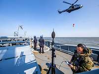 Ein Mann in Uniform steht auf einem Schiff neben einem Mikrofon, im Hintergrund ein Hubschrauber.
