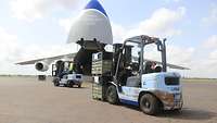 Zwei Gabelstapler mit Transportkisten fahren auf dem Rollfeld zu einem Transportflugzeug mit geöffneter Bugklappe hin.