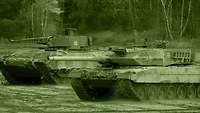 Ein Kampfpanzer fährt auf einer Freifläche neben einem Schützenpanzer.