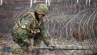 Ein Soldat hockt an einem Zaun aus Stacheldraht und schiebt eine Metallstange zwischen die Drähte.