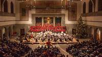 Festliche Konzertsituation in einer weihnachtlich geschmückten Kirche. Im Vordergrund Musiker, im Hintergrund ein Chor.