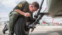 Ein Soldat in Fliegeroverall mit Werkzeugen beugt sich am Boden über das Rad eines Hubschraubers.